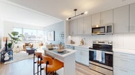 This thoughtfully designed single-floor residence showcases a cohesive modern aesthetic throughout its open-concept layout. The kitchen is a standout feature, equipped with stainless steel appliances including a range, microwave, dishwasher, and full-size refrigerator. Soft grey cabinetry extends to the ceiling, complemented by white subway tile backsplash and clean white countertops. A peninsula with seating for two provides a casual dining option and serves as the natural divide between the kitchen and living area. Light hardwood flooring runs throughout, adding warmth to the contemporary palette. The living room is bathed in natural light from large windows that frame open city views, creating a bright and airy atmosphere. A soft blue accent wall adds a subtle touch of color to the otherwise neutral space. The bedroom continues the light-filled theme with its own large window offering city views, sage green accent walls, and ample closet storage. The bathroom features large-format tile flooring, a white vanity with under-sink storage, and a well-lit mirror. Overall, the residence presents a clean, modern design with carefully selected finishes that create a cohesive and inviting living environment.