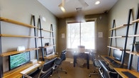 This single-floor workspace is thoughtfully designed to accommodate multiple users in a compact yet organized environment. The room features olive-toned walls that create a calm, focused atmosphere, complemented by tile flooring that adds a clean, professional aesthetic. Natural light streams through a centrally positioned window fitted with a white blind, illuminating the space and offering a view of surrounding greenery outdoors.

Three workstations are arranged throughout the room, each equipped with ergonomic black rolling chairs that prioritize comfort during extended use. A standout feature is the lean-style, black metal and wood shelving unit along the left wall, which provides tiered storage and display space, including a desktop computer setup and small decorative plant. A dedicated printer station occupies the center of the room, positioned conveniently for shared access. Additional desk surfaces along the right wall offer further workspace options.

Overhead, a ceiling fan ensures air circulation, while recessed lighting supplements the natural light. The overall design balances functionality with a modern, minimalist aesthetic, making this a practical shared workspace suited for focused tasks and general office use.