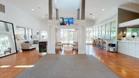 The clubhouse at Emerald Pointe in Hollywood, Florida is a thoughtfully designed single-floor community facility that combines functionality with a warm, inviting atmosphere. The expansive main hall greets visitors with soaring ceilings, warm terracotta tile flooring, and an abundance of natural light streaming through floor-to-ceiling windows and glass walls that offer views of the outdoor pool area. Prominent structural columns frame the open interior, adding architectural character throughout the space.

The welcoming entrance features elegant accent seating with ornate high-back chairs positioned near a staffed reception desk, creating a polished first impression. A comfortable lounge area anchors the center of the main hall, furnished with neutral-toned seating arrangements centered around a round white coffee table, ideal for casual gatherings or quiet relaxation.

Adjacent to the main lounge, a well-equipped business center lines one side of the hall, offering computer workstations with natural light views — perfect for residents working remotely. A compact, neatly appointed kitchenette with white cabinetry, a breakfast bar, pendant lighting, and stainless steel appliances supports community events and everyday convenience. Decorative artwork and carefully placed greenery throughout enhance the bright, Florida-inspired aesthetic of the entire space.