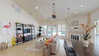 This single-story space functions as a well-appointed communal gathering area, thoughtfully organized into distinct zones that flow seamlessly into one another. The centerpiece is a generously proportioned lounge area anchored by soaring vaulted ceilings that amplify the sense of openness and airiness throughout. Recessed lighting and a ceiling fan complement the abundant natural light streaming through large windows and a set of French doors that open directly to an outdoor pool area, providing an inviting indoor-outdoor connection.

The lounge is furnished with a spacious sectional sofa, glass-topped coffee tables, and a media console, while a tile-surround fireplace adds a refined architectural focal point. Light-toned ceramic tile flooring runs throughout the entire space, enhancing the bright, clean aesthetic.

Adjacent to the lounge, a dedicated dining area features a dark wood table with upholstered chairs positioned beside oversized windows fitted with horizontal blinds, offering views of the surrounding landscape.

The refreshment kitchen is equipped with rich wood cabinetry with glass-front uppers, granite countertops, stainless steel appliances, and a beadboard-paneled peninsula with bar seating beneath pendant lighting — ideal for serving gatherings. A clearly marked refreshments room with a French-style glass door provides additional utility storage nearby.