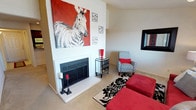 This single-story space in Arlington showcases a welcoming living area anchored by a white-framed fireplace, which serves as the room's focal point. The fireplace is flanked by decorative candleholders and topped with a striking oversized painting of a zebra on a bold red background, lending the space a dramatic and artistic character. The walls rise generously above the fireplace, emphasizing the vertical scale of the room and providing ample display space for wall art and décor.

The living area features neutral-toned carpeting throughout, providing a soft foundation that complements the vivid red and black color scheme carried through the furnishings and accessories. Seating includes plush red ottomans and a patterned accent chair in grey and white, accented with a red throw pillow. A mirrored side table adds a reflective, elegant touch beside the seating arrangement. A bold black-and-white damask area rug defines the sitting area and adds graphic contrast to the space.

A framed mirror on the adjacent wall enhances the sense of depth and light. Overall, the space presents a cohesive, energetic aesthetic defined by strong color contrasts and carefully curated decorative elements.