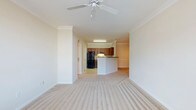 This single-floor residence is designed around an open-concept floor plan that seamlessly connects the living area to the kitchen, creating a spacious and cohesive living environment. The living space is finished with plush, neutral-toned wall-to-wall carpeting that adds warmth underfoot, while crisp white walls and elegant crown moulding lend a polished, classic character throughout.

A ceiling fan with a decorative medallion is centrally positioned in the living area, providing both functionality and a subtle architectural accent. The smooth, textured ceiling complements the clean lines of the moulding, reinforcing the refined aesthetic of the space.

The kitchen is situated at the rear of the open layout and features a raised peninsula counter with a white base, ideal for casual dining or meal preparation. Medium-toned wood cabinetry lines the kitchen walls, paired with a black refrigerator and matching appliances. An arched passageway adds an architectural detail that softens the transition between spaces. Recessed and under-cabinet lighting ensures the kitchen remains well-lit and practical. The overall ambiance is bright, clean, and move-in ready.
