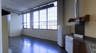 This single-floor space embodies a distinctive industrial character, drawing on its raw architectural roots to create an open and functional environment. Expansive factory-style windows span the width of the main living area, flooding the interior with abundant natural light and offering views of the surrounding urban streetscape. The large, multi-pane window grid is a standout architectural feature, lending the space a loft-like, warehouse-inspired aesthetic.

The flooring throughout is polished concrete, reflecting light and adding to the spacious, open feel of the interior. Exposed ductwork runs along the ceiling, reinforcing the industrial design language and contributing to the overall sense of volume and height.

A kitchen area is positioned to one side of the open floor plan, equipped with a freestanding electric range, a full-sized refrigerator, and cabinetry for storage. A range hood is mounted above the cooking area, providing practical ventilation. The kitchen integrates seamlessly into the open layout without visual separation, maintaining the airy, unobstructed feel of the space.

Overall, the space strikes a balance between utilitarian industrial design and livable comfort, making it well-suited for those who appreciate open-plan living with bold architectural character and strong natural light.