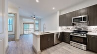Located at 3000 Hicks St in Houston, TX, this single-story unit showcases a thoughtfully designed open-concept layout with modern finishes throughout. The kitchen serves as a central highlight, featuring dark espresso cabinetry with sleek hardware, a textured stone-tile backsplash, white quartz countertops, and a large peninsula island with an integrated sink and stainless steel dishwasher. A full suite of stainless steel appliances, including a gas range, microwave, and French door refrigerator, completes the space.

The living area flows naturally from the kitchen and is finished with wood-look luxury vinyl plank flooring. Large sliding glass doors lead to a private balcony, allowing abundant natural light to fill the space. A ceiling fan adds both function and style.

The bedroom features plush carpet flooring, oversized windows that invite natural light, and direct access to a generously sized walk-in closet with built-in shelving and hanging rods. A second carpeted room offers flexible use as an additional bedroom or office.

The bathroom is appointed with gray wood-grain vanity cabinetry, white quartz countertops, dual mirrors, large-format tile flooring, and a glass-enclosed walk-in shower with floor-to-ceiling tile surround.