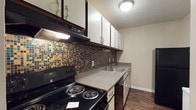 Located at 10612 Abercorn Extension in Savannah, Georgia, this single-floor unit offers a practical and well-appointed layout. The kitchen is a functional space equipped with a black electric range, built-in range hood, and a double-basin sink set into a continuous countertop. White upper cabinetry provides generous storage, while a striking mosaic tile backsplash in tones of blue, gold, and brown adds a decorative focal point. A textured ceiling and under-cabinet lighting complete the kitchen's design.

The main living area features rich hardwood-style flooring that extends throughout the unit, neutral warm-toned walls, and a textured ceiling. A full-width sliding glass door fitted with vertical blinds opens to a private balcony, allowing natural light to fill the room and providing views of the surrounding greenery.

The bedroom continues the cohesive flooring and neutral palette, with an arched architectural detail on one wall adding a subtle design element. A ceiling fan with a light fixture serves both the lighting and ventilation needs of the room. Clean white baseboards throughout the unit provide a crisp, finished look, contributing to the overall tidy and inviting character of this Savannah residence.