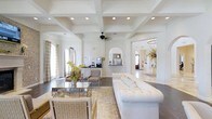 This expansive living area showcases a stunning coffered ceiling with recessed lighting, creating a sense of grandeur and depth. The space is characterized by its open floor plan, seamlessly connecting different functional areas. Arched doorways add architectural interest and lead to adjacent rooms, enhancing the flow of the space.

The room features a sophisticated color palette dominated by whites and neutrals, creating a bright and airy atmosphere. A plush white tufted sofa serves as a focal point, offering ample seating. The furniture arrangement includes modern chairs with woven leather details, complementing the contemporary glass coffee table.

Natural light floods the room through large windows, while sheer curtains provide a soft, diffused glow. The flooring appears to be a rich, dark hardwood, contrasting beautifully with the light-colored walls and furnishings. A small refreshment area with cabinetry is visible, suggesting convenience and functionality.

Decorative elements, such as a stylish ceiling fan and carefully chosen accent pieces, add character to the space. The overall design strikes a balance between comfort and elegance, creating an inviting environment perfect for both relaxation and entertaining.