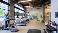 This fitness center offers a thoughtfully designed workout environment within a single-level space. Large floor-to-ceiling windows along one wall flood the interior with abundant natural light while providing views of the surrounding urban streetscape, creating an energizing atmosphere for exercise. The industrial-style ceiling exposes ductwork and structural elements, lending the space a contemporary aesthetic complemented by suspended linear lighting fixtures.

The cardio zone is prominently featured, housing a row of treadmills and a stationary bike, all positioned to take advantage of the natural light from the expansive windows. A wall-mounted flat-screen television is conveniently placed for entertainment during workouts. Full-length mirrors line one wall, visually expanding the space and allowing users to monitor their form during exercise.

The functional training area includes stability balls in multiple sizes, dumbbells, and exercise mats, catering to a variety of workout styles. The light-toned flooring throughout provides a clean, neutral foundation that complements the overall modern design. The open layout ensures ample room to move freely between equipment stations, making the space accessible and practical for diverse fitness routines.