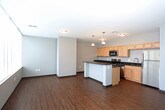 This single-floor unit presents a thoughtfully designed layout that balances open communal areas with more private living spaces. The standout feature is the expansive living area, where a wall of floor-to-ceiling windows fitted with horizontal blinds floods the room with natural light while framing striking panoramic city skyline views. Rich hardwood flooring runs throughout the main living and kitchen areas, adding warmth to the neutral gray and beige walls.

The kitchen is centrally positioned and well-appointed, featuring light maple cabinetry, dark granite countertops, and a full suite of stainless steel appliances, including a refrigerator, range, microwave, and dishwasher. A kitchen island with pendant lighting overhead provides additional prep space and serves as a natural divider between the kitchen and the open living area.

The bedrooms are carpeted in a deep charcoal tone, each offering a quiet, comfortable atmosphere with ceiling fans and simple, neutral walls. The bathroom is finished with warm hardwood-style flooring, a single-basin vanity with granite countertops and wood cabinetry, and a corner walk-in shower with a glass enclosure. Overall, the unit presents a clean, functional aesthetic with modern finishes throughout.