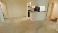 This single-floor residence offers a practical and comfortable layout centered around an open-concept living and kitchen area. The living space is finished with wall-to-wall carpeting in a warm neutral tone, creating a soft and inviting atmosphere throughout. A peninsula counter separates the kitchen from the main living area, providing both a functional food preparation surface with dark granite-style countertops and a natural visual boundary between spaces. The kitchen is equipped with black appliances, including a gas range and a refrigerator with a water dispenser, complemented by light wood cabinetry that adds a warm, understated aesthetic. Under-cabinet ventilation above the range highlights the kitchen's functional design. The walls throughout are painted in a soft, muted tone that enhances the sense of openness and keeps the space feeling bright and airy. The layout allows for easy flow between the living and kitchen areas, making the space well-suited for everyday living. The open sightlines and seamless transition between rooms contribute to an overall sense of spaciousness and ease of movement throughout the home.