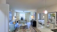 This thoughtfully designed single-floor residence offers an open and airy layout that seamlessly connects the living, dining, and sleeping areas. The living room serves as the heart of the space, furnished with a light blue sofa, a metallic coffee table, and a vibrant patterned area rug that adds warmth and texture to the light hardwood flooring. A bold piece of wall art anchors the media console, which houses a large flat-screen television, creating a stylish focal point.

Natural light floods the space through multiple windows dressed with elegant gold and white patterned curtains, enhancing the bright and welcoming ambiance. A ceiling fan with a integrated light fixture keeps the space comfortable. Adjacent to the living area, a glass-topped desk and chair create a functional workspace bathed in natural light.

The bedroom, visible through an open doorway, is neatly appointed with a crisp white bed and matching bedside lamps, offering a calm and restful retreat. The kitchen area, partially visible in the foreground, features attractive marble-look countertops that contribute a refined, upscale touch. Throughout the space, the cohesive blend of contemporary furnishings and warm accents creates a polished, inviting atmosphere.