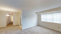 This single-floor space presents a straightforward, functional layout with a clean and neutral aesthetic throughout. The main living area is generously proportioned, finished with wall-to-wall carpet in a light beige tone that contributes to the room's warm, understated atmosphere. Walls are painted in crisp white and soft cream, lending a bright and airy feel to the interior.

A large window fitted with vertical blinds anchors one wall of the living area, allowing natural light to fill the space while offering adjustable privacy. The ceiling is smooth and standard height, maintaining a consistent, uncluttered look throughout.

An open passageway connects the living area to an adjacent kitchen space, which is partially visible from the main room. A white refrigerator can be seen within, suggesting a practical, equipped kitchen layout. The transition between areas is defined by a subtle architectural partition, creating a sense of separation without fully closing off the spaces.

Overall, the unit offers a clean, move-in-ready environment with a simple, no-frills design that prioritizes functionality and ease of use.