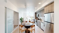 This thoughtfully designed single-floor residence offers a contemporary open-concept layout that seamlessly connects the kitchen, dining, and living areas. The kitchen is outfitted with wood-toned cabinetry, white tile backsplash, stainless steel appliances including a refrigerator, electric range, microwave, and dishwasher, and light countertops that give the space a clean, modern aesthetic. Light hardwood flooring runs throughout, enhancing the sense of continuity and warmth.

The dining area accommodates a round table with seating for multiple guests, anchored beneath a track lighting fixture. The living area is bathed in natural light from a large corner window offering open street-level views of a vibrant neighborhood, with tree-lined streets visible from within. A secondary lounge or flex space features a comfortable sofa and wall art, providing a relaxed retreat.

The bedroom is bright and airy, centered around a large window framing charming neighborhood views, creating a peaceful and connected atmosphere. The bathroom is clean and functional, featuring a full soaking tub with shower combination, white tile surround, and wood-accented vanity cabinetry. Throughout the residence, white walls and abundant natural light contribute to an open, inviting ambiance suited for modern everyday living.