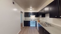 This single-story unit at Royal Arms offers a well-organized layout with distinct living, sleeping, and functional zones clearly defined throughout the floor plan. The kitchen serves as a standout space, featuring bold dark cabinetry spanning both upper and lower sections, light-toned countertops, wood-style plank flooring, and white appliances including an electric range and dishwasher. A double stainless steel sink sits beneath ample counter space, providing practical utility for daily use. Crown molding along the ceiling adds a refined finishing touch to the kitchen area.

The living area is carpeted and features a large sliding glass door fitted with vertical blinds, inviting natural light and offering views of the surrounding greenery. Multiple bedrooms continue the carpeted flooring theme, with neutral warm-toned walls contributing to a calm, understated aesthetic. One bedroom features crown molding and a ceiling fan with integrated lighting, while windows with blinds allow for natural light and privacy control throughout the sleeping areas.

The bathroom is well-appointed with a wide vanity, extended countertop, large mirror with overhead lighting, white cabinetry, and a glass-enclosed walk-in shower, creating a clean and functional space.