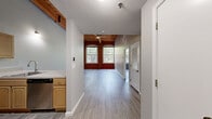 This one-bedroom, one-bathroom loft-style unit showcases a compelling mix of industrial heritage and contemporary design across a single, well-proportioned floor. The open-plan living and kitchen area is immediately striking, with soaring ceilings clad in warm timber planks supported by heavy exposed wooden beams and industrial-style pipe installations overhead. Light gray walls and light-toned hardwood flooring run throughout, creating a bright and cohesive aesthetic.

The kitchen is thoughtfully equipped with light maple cabinetry, stone-effect countertops, stainless steel appliances including a refrigerator, electric range, and dishwasher, as well as a stainless steel sink. A breakfast bar peninsula connects the kitchen to the living area, making the space ideal for both cooking and entertaining.

The bedroom is a comfortable, well-lit room featuring the same hardwood flooring and timber beam ceiling detail, with a large multi-pane window framing views of surrounding greenery and allowing generous natural light to fill the space. The exposed brick wall adds a defining architectural character to this room.

The bathroom is finished with large-format tile flooring, a full-size tub-shower combination, a toilet, and a maple vanity with a countertop sink, complemented by a wall-mounted mirror and overhead lighting fixture. The overall ambiance of the unit is urban yet warm, with a strong industrial-chic character.