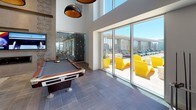 This thoughtfully designed single-floor amenity space combines modern aesthetics with a full suite of social and recreational features. The gourmet kitchen is a standout, showcasing bold hexagonal mosaic tile walls, warm wood cabinetry, dark stone countertops, and professional-grade stainless steel appliances including a gas range. A generous marble-topped island with vibrant yellow bar stools and globe pendant lighting serves as both a functional workspace and a social gathering point, with floor-to-ceiling windows framing sweeping city views beyond.

The lounge area offers a welcoming atmosphere with soaring ceilings, a linear gas fireplace set within a textured tile surround, a wall-mounted television, built-in bookshelves, and a curated mix of contemporary furnishings accented by bold artwork. Adjacent to the lounge, a billiards room with double-height ceilings and large-format floor tiles opens directly to the outdoor terrace through expansive glass sliding doors.

The outdoor space features a rooftop pool deck with lounge chairs, bold yellow accent seating, and panoramic city skyline views. Throughout, a cohesive design palette of warm wood tones, grey tile, white walls, and yellow accents creates an energetic yet refined ambiance ideal for both relaxation and entertainment.
