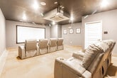 Located at 1555 Main St in Rahway, NJ, this dedicated screening room delivers an immersive cinematic experience within a compact, well-appointed single-level space. The room is designed with a clear focus on comfort and functionality, anchored by a large, wall-mounted projection screen at the front that commands immediate attention upon entry.

Seating is arranged in two rows of plush gray leather recliner chairs, each equipped with built-in cup holders — a thoughtful detail that enhances the theater atmosphere. The tiered layout ensures unobstructed sightlines from every seat. Decorative number placards mounted on the slate-blue walls add a charming, classic movie-house aesthetic.

The dark, matte-black ceiling is a defining architectural feature, contributing to the room's light-controlled environment while housing recessed spotlights and a ceiling-mounted projector. A cassette-style HVAC unit is integrated flush into the ceiling, ensuring climate comfort without compromising the visual design. Neutral-toned carpet tiles cover the floor, adding warmth and sound absorption.

A paneled entry door and a smart control panel on the wall provide easy access and room management. The overall ambiance is intimate yet polished, blending the feel of a private cinema with the refined finishes of an upscale amenity space.