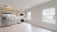 Located at 181 North Main Street in Suffolk, Virginia, this thoughtfully designed 2-bedroom, 1-bathroom residence offers a clean, modern aesthetic throughout. The open-concept living and kitchen area is bathed in natural light, courtesy of multiple double-hung windows that frame views of the surrounding city. The kitchen is outfitted with stainless steel appliances — including a range, dishwasher, refrigerator, and microwave — complemented by gray shaker-style cabinetry, a herringbone tile backsplash, and recessed lighting overhead. Light-toned flooring flows seamlessly across the entire living space, reinforcing the bright and airy atmosphere. The spacious primary bedroom is a standout, featuring three large windows on two walls that flood the room with natural light and provide sweeping outdoor views, along with a ceiling fan for added comfort. The second bedroom offers similar light-filled character. The bathroom is finished with white cabinetry, a single-sink vanity with brushed gold hardware, a tub-shower combination, and tile flooring. Entry to the unit is secured with a keypad-enabled smart lock, adding a modern convenience. The overall design palette — soft whites, warm neutrals, and brushed gold accents — creates a cohesive and inviting living environment.