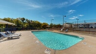 Located at 5000 Denton Hwy in Haltom City, Texas, this outdoor swimming pool area serves as a central recreational amenity for the two-story residential community. The generously sized pool features clearly marked depth indicators, ranging from a shallow entry point to deeper waters, and is equipped with metal entry railings for safe and easy access. The surrounding concrete sundeck provides ample space for lounging, complemented by poolside chaise lounge chairs and a shaded umbrella seating area, ideal for relaxing on warm Texas days. The entire pool area is enclosed by a black iron security fence with a gated entry, ensuring privacy and safety for residents. Tall light poles positioned throughout the enclosure extend the usability of the space into the evening hours. The backdrop of mature trees lining the perimeter adds a natural, shaded canopy and a sense of seclusion. Views of the two-story stucco residential buildings are visible from the pool deck, reinforcing the close-knit community feel. A secondary pool area is also present on the property, similarly fenced and surrounded by manicured landscaping. The open-sky setting under the clear Texas blue sky creates a bright, inviting atmosphere throughout the entire outdoor space.