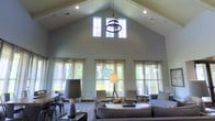 This single-story space makes a bold architectural statement with its dramatic vaulted cathedral ceiling, which rises to an impressive peak and creates an expansive, airy atmosphere throughout the main living area. A row of tall clerestory windows at the apex of the gable floods the space with natural light from above, complementing the generous arrangement of full-height windows along the surrounding walls that offer lush, verdant outdoor views.

The living area is thoughtfully appointed with neutral-toned upholstered seating, including a plush sofa and a pair of accent chairs, arranged around a central coffee table to encourage comfortable conversation. A decorative wrought-iron chandelier suspended from the ceiling peak serves as a focal point, while coordinating table and floor lamps provide warm, layered lighting throughout.

Sheer drapery panels in soft, neutral hues line the windows, softening the abundant natural light while maintaining an open, welcoming feel. The overall color palette — composed of muted greens, warm grays, and earthy tones — lends the space a calm, sophisticated ambiance. The combination of soaring architecture, natural light, and carefully curated furnishings results in an elegant yet inviting environment.