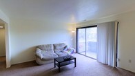 This single-floor residential space offers a clean, functional layout centered around a welcoming living area. The living room is finished with wall-to-wall carpeting that adds warmth underfoot, while smooth, lightly textured ceilings contribute to the overall sense of calm and simplicity throughout the space. Natural light is a defining feature of the room, streaming in generously through a large sliding glass door fitted with vertical blinds. The sliding door opens directly onto a balcony, providing an easy indoor-outdoor connection and a view of the surrounding exterior environment. The living area comfortably accommodates a sofa and coffee table arrangement, making it a practical and inviting space for everyday living. The neutral color palette of soft whites and warm beiges creates a versatile backdrop well-suited to a variety of furnishing styles. The overall ambiance is modest and understated, with a straightforward design that prioritizes comfort and livability. The balcony access adds an appealing outdoor dimension to the space, enhancing both ventilation and natural light throughout the day.