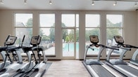 Located at 100 Northwoods Dr in Cary, NC, this single-floor fitness center is a well-equipped facility designed to support a comprehensive workout experience. The space is bright and open, characterized by light wood-look flooring, neutral walls, and an energizing orange accent column that adds a bold visual focal point throughout the gym floor.

The cardio zone is positioned along a wall of large, floor-to-ceiling windows and a glass door that opens directly to an outdoor pool and deck area, allowing natural light to flood the space and offering motivating views of the lush greenery beyond. Treadmills and elliptical machines are arranged to take full advantage of this scenic outlook. Track lighting runs throughout the ceiling, ensuring consistent illumination across all workout areas.

The main fitness floor accommodates a row of treadmills, a set of cycling bikes, and a variety of strength training machines including cable systems, weight benches, and a dumbbell rack. Arched transom windows near the roofline add architectural character and bring in additional natural light. Full-length mirrors are positioned to assist with form and enhance the sense of space.

A dedicated restroom with ADA-compliant grab bars and wood-look tile flooring is conveniently accessible within the facility. The open layout and thoughtful zoning make this fitness center functional, accessible, and inviting for users of all fitness levels.