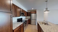 This single-floor residence offers a thoughtfully designed layout that balances practicality and comfort throughout its living spaces. The kitchen serves as a functional centerpiece, featuring rich dark-wood cabinetry with brushed hardware, light granite-style countertops, and stainless steel appliances including a refrigerator, over-the-range microwave, and electric range. Light hardwood-style flooring runs through the kitchen and connects seamlessly to the entry and living areas, enhancing the sense of continuity. The open living area benefits from abundant natural light, courtesy of large windows fitted with vertical blinds and a sliding glass door, creating a bright and airy atmosphere. The bedrooms are generously proportioned, each finished with soft neutral carpeting and sizable windows with horizontal blinds that invite natural light while maintaining privacy. One bedroom features a double-door closet, offering practical storage. The bathrooms are clean and functional; one includes a soaking tub with shower combination and tiled flooring, while the other features a vanity with dark wood cabinetry, a large mirror, and matching tile floors. Throughout, the neutral color palette and consistent finishes create a cohesive, move-in-ready environment.