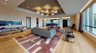 This single-floor café is a thoughtfully designed, open-plan space that seamlessly blends lounge, dining, and service areas into a cohesive, contemporary environment. The layout is distinguished by a striking contrast of wide-plank hardwood flooring in the lounge zone and mixed tile patterns in the entry and transition areas, grounding each section with its own distinct character.

Floor-to-ceiling windows along one full wall flood the interior with natural light while offering panoramic views of the surrounding urban skyline and waterfront. Sheer drape curtains soften the light and add an element of refinement. The lounge area is furnished with leather and upholstered sofas, armchairs, and glass coffee tables atop a bold geometric area rug, centered beneath a sculptural pendant chandelier with exposed red wiring—a deliberate design accent.

A dedicated dining area features communal tables and bar-height seating, positioned adjacent to a sleek service counter equipped with coffee and beverage equipment. Large-scale photographic and artistic wall murals serve as dramatic focal points throughout the space.

Glass doors open directly onto an outdoor terrace, extending the usable area and reinforcing the connection to the outdoors. The overall ambiance is sophisticated yet approachable, with curated furnishings, layered lighting, and artistic elements creating a dynamic and inviting atmosphere.