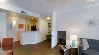 This single-story space showcases a thoughtfully designed open floor plan with hardwood-style flooring and bright white walls throughout. The living area flows naturally toward a pass-through kitchen counter, which opens into a warmly lit kitchen featuring recessed lighting, dark appliances, and ample counter space — ideal for both cooking and casual interaction with guests.

Adjacent to the living area, a built-in desk nook provides a dedicated workspace complete with overhead lighting and open shelving above, offering practical storage and display space. This alcove is cleverly integrated into the layout without interrupting the overall flow of the space.

A standout feature is the built-in pet retreat — a small, enclosed nook at floor level, thoughtfully designed to provide a cozy, designated area for a pet. Crown molding lines the ceilings, adding a refined architectural detail, while recessed and surface-mounted lighting ensures consistent illumination throughout. The overall ambiance is clean, functional, and well-organized, with each area of the space serving a distinct purpose while maintaining a cohesive, contemporary aesthetic.