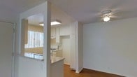 This single-floor space showcases an open-concept layout that seamlessly connects the kitchen, dining, and living areas. The kitchen is outfitted with white cabinetry extending to the ceiling, providing ample storage, and features a white gas range. A breakfast bar with a light countertop serves as a natural divider between the kitchen and the adjacent living space, offering both a casual dining option and an efficient use of space.

Warm hardwood-style flooring runs throughout the main living areas, adding a cohesive and inviting feel. The living area is equipped with a ceiling fan, contributing to the overall comfort of the space. Natural light filters in through vertical blinds on a nearby window, brightening the interior without overwhelming it.

The textured ceiling adds a subtle architectural detail throughout, while the clean white walls and trim create a neutral, versatile backdrop. A structural column near the kitchen adds character to the layout. Overall, the space is designed for practical, everyday living with a clean, functional aesthetic.