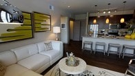 This single-floor space showcases an open-concept layout that connects the living area and kitchen in a cohesive, modern design. The living area features rich, dark hardwood flooring that extends throughout, anchoring a clean, contemporary aesthetic. A white sofa paired with decorative pillows sits opposite a round marble-top coffee table, while a bold yellow abstract artwork adds a vibrant focal point to the neutral gray walls. A table lamp provides warm ambient lighting, complementing the overall inviting atmosphere.

The kitchen is seamlessly integrated into the open floor plan and is outfitted with dark espresso cabinetry, stainless steel appliances, and a light-toned countertop that wraps around a peninsula-style breakfast bar. Two upholstered bar stools offer casual seating at the bar. A pendant light in an amber tone hangs above the kitchen area, adding warmth, while recessed track lighting along the ceiling ensures practical illumination throughout.

A hallway visible from the living area leads to additional rooms beyond, hinting at further private spaces. The overall ambiance is polished and contemporary, balancing neutral tones with selective accents for a refined, livable aesthetic.