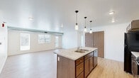 Located at 5102 Shelby Ave in Rapid City, South Dakota, this single-floor home offers four bedrooms and two full bathrooms within a well-designed, functional layout. The open-concept kitchen and living area serves as the heart of the home, anchored by a central island with a built-in sink and light granite-style countertops. Rich dark wood cabinetry lines the kitchen walls, complemented by black appliances including a refrigerator, range, and built-in microwave. Three pendant lights hang above the island, adding a modern touch to the space. Light hardwood flooring flows throughout the main living areas, creating a bright and cohesive feel. The spacious living room benefits from large windows and a glass exterior door that invite abundant natural light and provide outdoor views. Exposed ductwork along the ceiling adds an industrial design element. The bedrooms feature plush carpet flooring and windows overlooking the surrounding neighborhood. Closets include built-in shelving and hanging rods for organized storage. Both bathrooms are finished with granite-topped dark wood vanities and black hardware accents. The primary bathroom includes hexagonal tile flooring, a bathtub, and a dual-sink vanity. The secondary bathroom features light hardwood flooring and a clean, minimalist design.