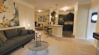This single-floor residence features an open-concept layout that integrates the living, dining, and kitchen areas into one cohesive, flowing space. The living area is anchored by plush carpeting and accommodates comfortable seating, with a round coffee table serving as a focal point. Crown molding runs throughout, adding a refined architectural detail to the warm, neutral-toned walls.

The kitchen is a standout feature, showcasing dark espresso cabinetry that contrasts sharply against light countertops, including a curved breakfast bar with stylish spiral bar stools beneath pendant lighting. The kitchen transitions to hardwood-style flooring, clearly delineating it from the carpeted living and dining areas. Black appliances complement the cabinetry, while a tile backsplash adds texture and depth.

The dining area sits just beyond the living space, offering an intimate setting with a table and chairs framed by soft, ambient lighting. Track lighting in the kitchen and pendant fixtures above the bar ensure the space is well-lit throughout. The overall ambiance is warm and inviting, balancing contemporary design elements with comfortable, livable details.