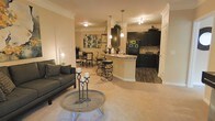 This single-floor residence features an open-concept layout that integrates the living, dining, and kitchen areas into one cohesive, flowing space. The living area is anchored by plush carpeting and accommodates comfortable seating, with a round coffee table serving as a focal point. Crown molding runs throughout, adding a refined architectural detail to the warm, neutral-toned walls.

The kitchen is a standout feature, showcasing dark espresso cabinetry that contrasts sharply against light countertops, including a curved breakfast bar with stylish spiral bar stools beneath pendant lighting. The kitchen transitions to hardwood-style flooring, clearly delineating it from the carpeted living and dining areas. Black appliances complement the cabinetry, while a tile backsplash adds texture and depth.

The dining area sits just beyond the living space, offering an intimate setting with a table and chairs framed by soft, ambient lighting. Track lighting in the kitchen and pendant fixtures above the bar ensure the space is well-lit throughout. The overall ambiance is warm and inviting, balancing contemporary design elements with comfortable, livable details.