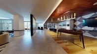 This expansive single-floor amenity space is thoughtfully designed to serve as a premier social and wellness hub. The interior immediately impresses with its warm wood-paneled ceilings, complemented by recessed and cove lighting that creates an inviting, upscale atmosphere throughout. Large floor-to-ceiling glass walls and windows flood the space with natural light while providing views of the surrounding exterior.

The layout is organized into distinct functional zones that flow seamlessly into one another. A generous lounge area features a curved sectional sofa, accent chairs, and artfully selected rugs atop light hardwood flooring, anchored by a striking geometric-patterned feature wall with bold pendant lighting overhead. Built-in shelving units provide curated display space, adding a library-like sophistication to one corner.

A dedicated kitchenette is finished with rich wood cabinetry, a marble-inspired backsplash, white quartz countertops, a stainless steel dishwasher, and a beverage cooler — ideal for entertaining. A bar-height counter with stools offers additional gathering space nearby.

The game area includes a foosball table beneath a dramatic chandelier, while a glass-enclosed fitness center with modern equipment is accessible directly from the main floor. Textured stone accent walls and an elevator add to the space's polished, contemporary character.