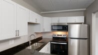 This single-floor unit offers a thoughtfully designed layout that balances functionality with modern finishes. The kitchen is a standout feature, equipped with stainless steel appliances including an electric range, over-the-range microwave, and refrigerator. Crisp white shaker-style cabinetry provides ample storage, while dark granite countertops add a striking contrast. A double stainless steel sink with a contemporary faucet completes the space. The kitchen opens to a spacious living and dining area finished with durable hardwood-style flooring in a warm grey tone, creating a seamless and cohesive feel. Natural light filters in through large windows fitted with vertical blinds, brightening the open-concept living space. Baseboard heating runs along the perimeter of the main living areas, ensuring comfortable warmth throughout. The bedrooms are carpeted in a neutral tan tone, offering a softer underfoot experience, and each room benefits from natural light via sizable windows. A hallway connects the living areas to the private rooms, maintaining a clear and organized flow. The bathroom features a full-size soaking tub and shower combination with clean white tile surround and polished chrome fixtures. Consistent tile drop ceilings and neutral grey walls tie the entire unit together with a clean, understated aesthetic.