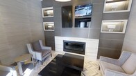 This contemporary lounge space showcases a thoughtfully designed interior with a strong emphasis on modern aesthetics and refined finishes. The focal point of the room is a striking feature wall clad in large-format gray tiles, housing a linear gas fireplace framed in crisp white stone surround. Above the fireplace, a wall-mounted flat-screen television is seamlessly integrated into the design, flanked by illuminated display niches that add both ambient lighting and architectural interest to the composition.

The feature wall's built-in alcoves are backlit, creating a warm glow that contrasts elegantly with the cooler gray tile palette. The seating arrangement includes classic wingback-style chairs upholstered in soft gray fabric, positioned symmetrically around a sleek, dark rectangular coffee table — offering a comfortable and balanced gathering area.

Underfoot, the herringbone-patterned flooring in a light, natural tone adds texture and visual movement to the space, complementing the monochromatic color scheme. The overall ambiance is polished and inviting, blending clean architectural lines with warm lighting accents. This space functions effectively as a communal lounge or reception area, combining functional design with an upscale, contemporary atmosphere.