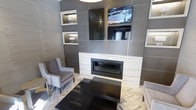 This contemporary lounge space showcases a thoughtfully designed interior with a strong emphasis on modern aesthetics and refined finishes. The focal point of the room is a striking feature wall clad in large-format gray tiles, housing a linear gas fireplace framed in crisp white stone surround. Above the fireplace, a wall-mounted flat-screen television is seamlessly integrated into the design, flanked by illuminated display niches that add both ambient lighting and architectural interest to the composition.

The feature wall's built-in alcoves are backlit, creating a warm glow that contrasts elegantly with the cooler gray tile palette. The seating arrangement includes classic wingback-style chairs upholstered in soft gray fabric, positioned symmetrically around a sleek, dark rectangular coffee table — offering a comfortable and balanced gathering area.

Underfoot, the herringbone-patterned flooring in a light, natural tone adds texture and visual movement to the space, complementing the monochromatic color scheme. The overall ambiance is polished and inviting, blending clean architectural lines with warm lighting accents. This space functions effectively as a communal lounge or reception area, combining functional design with an upscale, contemporary atmosphere.