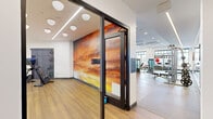 This expansive fitness center is thoughtfully designed to support a full range of workout disciplines across a single open floor. The main gym area features warm wood-look flooring contrasted with rubber matting beneath the strength equipment, and is bathed in natural light streaming through large, black-framed windows that offer views of the surrounding courtyard. The space houses a comprehensive selection of Matrix-brand strength machines, a multi-rack free weights station, battle ropes, a WaterRower, and an assortment of kettlebells and medicine balls. Cardio equipment, including treadmills, ellipticals, and stair climbers, is positioned near the windows to maximize the outdoor connection. A dedicated cycling studio features a striking floor-to-ceiling sunset mural, full-wall mirrors, stationary bikes with individual screens, and a mounted display — creating an immersive workout environment. An adjoining functional training room holds stability balls and additional accessories behind sleek glass-and-black-frame partitions. A built-in wooden shelving unit and a wall-mounted smart display provide both storage and digital connectivity. Exposed ductwork, recessed lighting, and ceiling fans add an industrial-modern touch to the bright, white interior, while the climbing wall adds a dynamic recreational element to the overall facility.