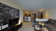 This single-floor residence offers an open-concept layout that seamlessly connects the living area and kitchen, creating a cohesive and functional living environment. The living area is anchored by a large flat-screen television mounted on a sleek media console, accompanied by a bold black-and-white urban cityscape wall mural that serves as a striking focal point. A plush grey sectional sofa and a glass-top coffee table with decorative books and fresh flowers add comfort and style to the space. Recessed lighting and track lighting illuminate the interior evenly, complementing the neutral toned carpet that runs throughout. The kitchen features warm wood cabinetry, stainless steel appliances including a microwave and range, and a white breakfast bar with metal bar stools, ideal for casual dining. A closet or storage area is visible near the entryway, which also includes a solid wood door providing access to the unit. The overall ambiance is modern and urban-inspired, with thoughtful design details that make the space feel both practical and inviting.