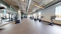 Located at 1624 W Babcock in Bozeman, Montana, this fitness center offers a generous, open-plan workout space spread across a single floor. The facility showcases a contemporary industrial aesthetic, with exposed concrete ceilings, prominent HVAC ductwork, and structural columns that lend the space a raw, modern character. Linear LED lighting runs throughout, ensuring consistent, well-distributed illumination across the entire floor.

The space is clearly divided into functional zones. The cardio area lines the windowed perimeter, where large windows flood the room with natural light and provide views of the surrounding Bozeman streetscape. Equipment includes treadmills, ellipticals, stationary bikes, and a rowing machine. The strength and free weights zone occupies the interior, featuring squat racks, weight benches, a full dumbbell rack, and cable machines, all reflected in full-length wall mirrors that enhance the sense of space. A dedicated functional training area houses a multi-attachment suspension and resistance band station alongside plyo boxes, supporting diverse workout modalities.

Durable, speckled rubber flooring covers the entire space, providing safety and resilience underfoot. The open layout allows for generous circulation and flexibility in equipment arrangement, making the facility well-suited for a wide range of fitness activities.