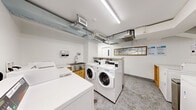 Located at 1420 Rue Crescent in Montréal, this shared laundry facility offers a well-organized, functional space designed to accommodate multiple users efficiently. The layout is divided into two connected areas: a main laundry room and an adjacent corridor, together providing a comfortable amount of operational space.

The main laundry room houses a substantial lineup of commercial-grade front-loading washers and top-loading dryers, all finished in white and arranged along the walls to maximize usability. A built-in utility sink with warm wood cabinetry below provides a practical handwashing or pre-treatment station. A dedicated folding counter offers additional convenience for users. A framed decorative mirror with an art-inspired design adds a subtle aesthetic touch to the otherwise utilitarian environment.

The connecting corridor features a sleek marble-topped bench, white walls, and framed artwork, contributing a clean and welcoming transitional feel. Exposed ventilation ductwork runs along the ceiling throughout, while bright linear fluorescent lighting ensures thorough illumination across both areas. Speckled terrazzo flooring runs continuously through the space, offering durability and a polished appearance. The facility is well-ventilated and neatly maintained, presenting a practical and accessible laundry solution.