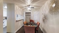 This single-story space includes a intimate dining area designed for comfortable everyday living. The dining room features dark hardwood-style flooring that grounds the space, complemented by smooth, neutral-toned walls with subtle angular detailing that adds visual interest. A ceiling fan with an integrated light fixture serves as the central overhead element, providing both air circulation and ambient lighting. A built-in pass-through shelf on one wall offers a functional connection to an adjacent area, ideal for serving or display. A closet with a standard rod provides convenient storage directly within the dining space. Wall décor, including a starburst accent piece, adds a stylish finishing touch. The room comfortably accommodates a round dining table with upholstered seating, creating a welcoming setting for meals. The overall ambiance is warm and understated, with a cohesive neutral color palette throughout.