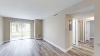 This single-floor unit has been thoughtfully updated with a clean, modern aesthetic carried consistently throughout every room. Light-toned, wood-look plank flooring runs across the entire space, complemented by crisp white baseboards and neutral warm-beige walls that create a bright, welcoming atmosphere.

The open living area is generously proportioned, featuring a sliding glass door that invites abundant natural light and offers views of lush greenery outside. A dedicated entry area includes a built-in electrical panel and provides a clear separation from the main living space. The layout flows naturally into a well-appointed galley-style kitchen, which features white shaker cabinetry with brushed nickel hardware, granite-patterned countertops, stainless steel appliances including a full-size refrigerator and electric range, and a window above the sink area providing additional natural light.

A secondary room, suitable as a bedroom or flex space, includes a window with blinds and recessed lighting, offering a quiet and versatile area. The bathroom is a standout feature, showcasing large-format marble-look tile in the walk-in shower, a granite-top vanity with an undermount sink, and brushed nickel fixtures that tie the design together with an upscale finish. Throughout the unit, HVAC vents and a central thermostat reflect a complete and functional infrastructure.