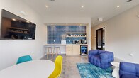 This single-story space presents a contemporary, well-appointed interior designed for comfort and functionality. The layout flows from an entry reception area—equipped with vibrant teal and yellow molded chairs and a wood-and-slate credenza—through glass-partitioned walls with sleek black frames that allow sightlines across the entire floor plate.

The main lounge area is a standout feature, anchored by a pair of plush, tufted blue sofas arranged on a patterned blue area rug. Large floor-to-ceiling sliding glass doors flood the space with natural light and provide direct views to the exterior, enhancing the open, airy atmosphere. Polished concrete floors extend throughout, contributing a clean, industrial-modern aesthetic.

A built-in beverage station serves as a focal point along one wall, clad in rich cobalt square tiles with illuminated open shelving, light wood cabinetry, a countertop bar with stools, a beverage refrigerator, and a coffee station. A wall-mounted flat-screen television is positioned nearby, complementing the casual lounge seating.

A long corridor connects the main areas to additional rooms, maintaining the bright, white palette and polished floors, and leading toward glazed exterior doors at the far end.