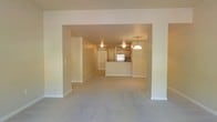 This single-floor space is designed with an open-concept layout that promotes a natural flow between the living area and the kitchen. A wide arched passageway connects the two main zones, creating a sense of openness and continuity throughout the interior.

The living area features wall-to-wall carpet in a neutral beige tone, contributing to a warm and understated ambiance. The soft, muted color palette of the walls complements the flooring, lending the space a clean and cohesive aesthetic.

The kitchen is positioned at the rear of the open plan and is anchored by a raised breakfast bar or peninsula, which serves as both a functional workspace and a subtle divider between the kitchen and the adjacent dining or living zone. Warm wood-toned cabinetry lines the kitchen walls, adding a natural element to the interior. A stainless steel appliance — likely a refrigerator — is visible, adding a modern contrast to the traditional cabinetry. A pendant light hangs above the kitchen area, providing focused illumination and a decorative focal point.

The layout also includes a hallway or secondary corridor visible to the left, suggesting additional rooms beyond the main living space. Overall, the space presents a functional, comfortable, and well-connected floor plan with a consistent and inviting neutral design throughout.