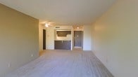 This single-floor unit offers an open and functional layout designed for everyday living. The main living area is spacious and bright, featuring neutral-toned carpeting that creates a warm and inviting atmosphere. The space flows naturally toward a dedicated kitchen area, which is separated by a tiled peninsula counter that doubles as a casual dining or prep surface. The kitchen is equipped with wood-toned cabinetry, a range, and an overhead microwave or ventilation unit, providing all the essentials in a compact and efficient arrangement. Tile flooring in the kitchen area offers a clean contrast to the carpeted living space. A doorway visible from the main room leads to additional areas of the unit, suggesting a well-organized floor plan. The ceilings feature a subtle texture finish with flush-mounted lighting, and HVAC vents throughout indicate a central air and heating system. Neutral wall tones throughout create a versatile backdrop suitable for a variety of interior styles. Overall, this unit presents a straightforward and practical living environment with a cohesive open-concept design connecting the key living and cooking spaces.