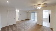 This single-floor space centers around a spacious main living area finished with wall-to-wall beige carpeting that extends throughout, contributing to a warm and comfortable atmosphere. The room is equipped with a ceiling fan fitted with an integrated light fixture, providing both air circulation and overhead lighting. A large sliding glass door dominates one wall, dressed with vertical blinds that allow for adjustable natural light and potential outdoor access. An HVAC wall unit is positioned beneath the sliding door, ensuring climate control within the space. The walls are painted in a clean, neutral white, lending a bright and open feel to the interior. A thermostat is mounted on one wall, offering convenient temperature management. A doorway on the left side of the room leads to an adjacent hallway, which appears to connect to additional areas of the space, including what looks like a closet with louvered doors visible in the background. The transition from carpet to tile flooring near the right edge suggests the presence of a separate room, likely a kitchen or entryway. Overall, the space presents a functional and straightforward layout with practical features throughout.
