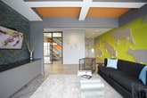 This dynamic single-floor commercial space is defined by its bold, creative design aesthetic and thoughtfully arranged zones for work, collaboration, and relaxation. The interior immediately captures attention with vivid, large-scale murals — including a striking multicolored chevron wall and a yellow accent wall adorned with dimensional animal silhouettes — infusing the space with energy and personality throughout.

A glass-enclosed conference room with black-framed partitions sits at the heart of the space, offering visual transparency while maintaining a defined meeting area. Clear acrylic chairs and a round wooden table create a modern, airy feel within. Adjacent to it, a casual lounge area with lime green accent chairs provides an informal setting for conversations or quick breaks.

Floor-to-ceiling windows along one exterior wall flood the space with abundant natural light and offer open views of the surrounding streetscape, creating a strong connection to the outdoors. Wood-look tile flooring flows continuously throughout, lending warmth and cohesion to the varied zones.

A communal dining or break area features sleek pendant lighting, a long white table with ghost chairs, and a beverage station set against a bold red ceiling accent. A quieter lounge nook offers a clean white bench seat for moments of focused retreat. The overall ambiance is modern, energetic, and purposefully designed for a creative, collaborative environment.