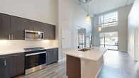 Located at 1774 Metro Avenue in Chula Vista, CA, this two-bedroom, two-bathroom residence spans 1,060 square feet across two floors. The main floor (820 sq ft) showcases soaring double-height ceilings and durable wood-look plank flooring throughout, creating a bright, open atmosphere. The entrance features floor-to-ceiling black-framed glass doors that frame street views and flood the space with natural light. The open-concept kitchen and living area is a standout, boasting dark-toned cabinetry extending to the ceiling, a white tile backsplash, light quartz countertops, a kitchen island, and a full suite of stainless steel appliances including a French door refrigerator, gas range, and built-in microwave. Hallways connect two well-sized bedrooms, including a 140 sq ft primary bedroom, each complemented by dedicated closets. Both bathrooms feature tub-shower combinations with white tile surrounds, chrome fixtures, and wood-look flooring. The upper floor (240 sq ft) is accessible via a staircase and offers additional living or bedroom space. Neutral warm-gray walls and consistent flooring unify the home's clean, contemporary aesthetic throughout.