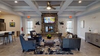 Located at 15 Old Carriage Rd in West Warwick, Rhode Island, this single-floor community amenity space is thoughtfully designed to offer residents a range of comfortable and functional areas. The centerpiece of the space is a generous lounge featuring a striking floor-to-ceiling stone fireplace, a wall-mounted TV, and elegant coffered ceilings with a ceiling fan. The seating area is furnished with leather sofas, accent chairs, and a patterned area rug over stylish tile flooring, creating a warm and inviting atmosphere. Natural light filters in through multiple windows, enhancing the open feel of the room. Adjacent to the lounge is a fully equipped kitchen featuring deep navy shaker-style cabinetry, marble-look countertops, stainless steel appliances including a refrigerator, range, microwave, and dishwasher, along with a coffee station. The business center offers a dedicated workspace with a wood-plank accent wall, built-in desks, desktop computers, and natural light from a side window. The fitness center is well-outfitted with treadmills, an elliptical, a cable machine, free weights, and rubber flooring, spread across two connected areas with coffered ceilings and ceiling fans throughout.